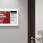 un pannello e-paper Sync-Sign montato sulla parete, accanto alla porta di una sala riunioni moderna. Lo schermo mostra in verde che la sala è disponibile.