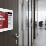 Una moderna targa porta ufficio digitale montata su una porta di legno scuro, con un corridoio d'ufficio sfocato sullo sfondo e persone che camminano.